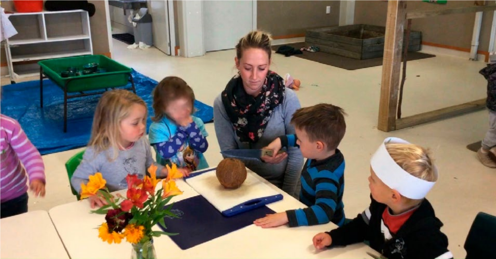 A photo of four children sitting or standing at a table. A teacher is kneeling next to one of the boys and holding his hand. He is holding a knife and is attempting to cut open a coconut which is sitting on the table