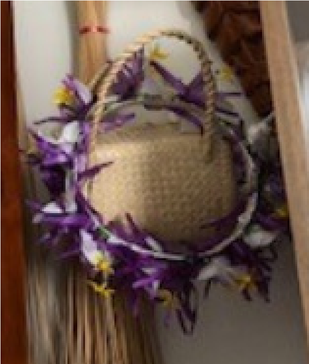 The picture displays a Cook Island 'ei katu (headpiece). It's mainly made of woven flax-like material, and has purple, yellow and white flowers or cloth around the brim